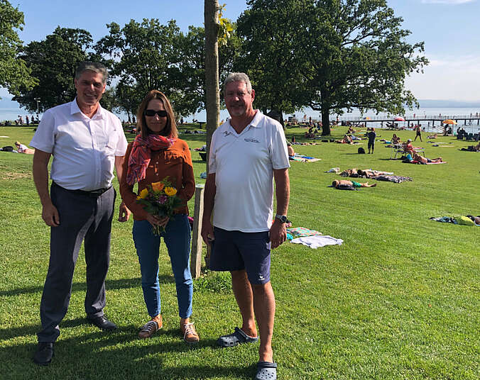 Zwei Männer und eine Frau mit Blumen in der Hand stehen auf der Liegewiese im Strandbad. Im Hintergrund der See.