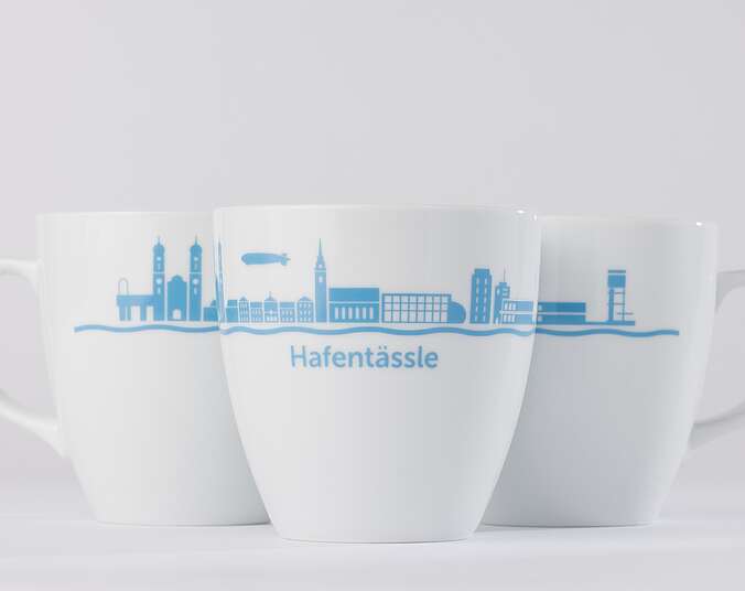 Drei weiße Tassen mit einer Silhouette von Friedrichshafen und dem Schriftzug Hafentaessle.
