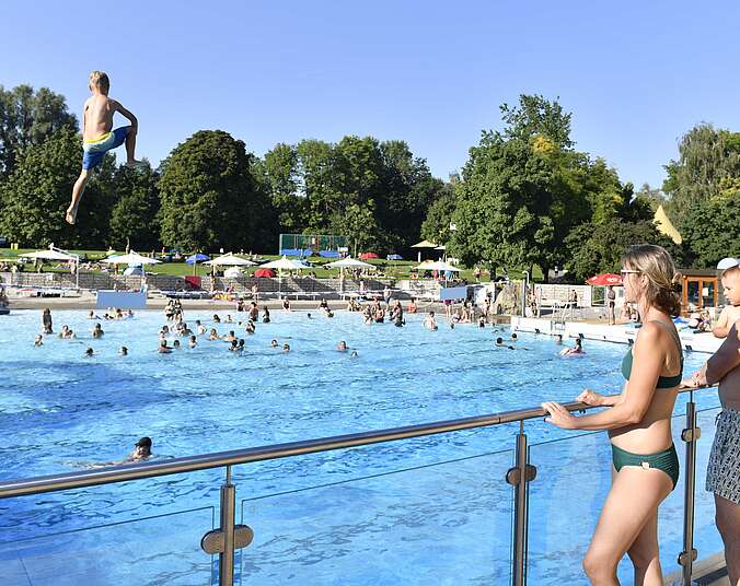 Zwei Personen stehen am Beckenrand und schauen aufs blaue Wasser, in dem Menschen schwimmen